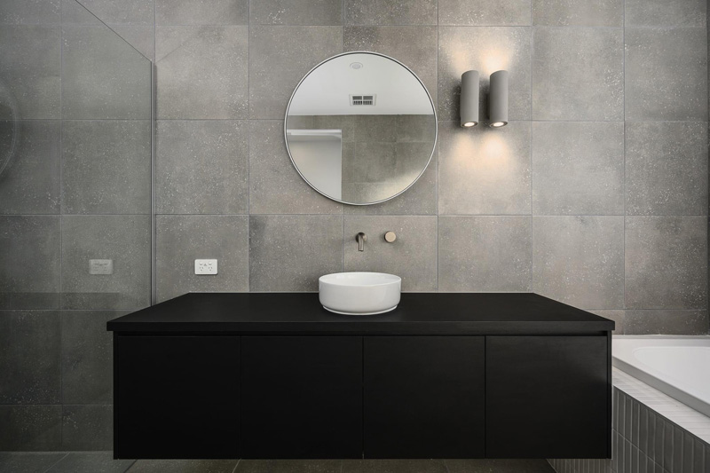 Grey tiled bathroom with a black vanity, round mirror and wall lights